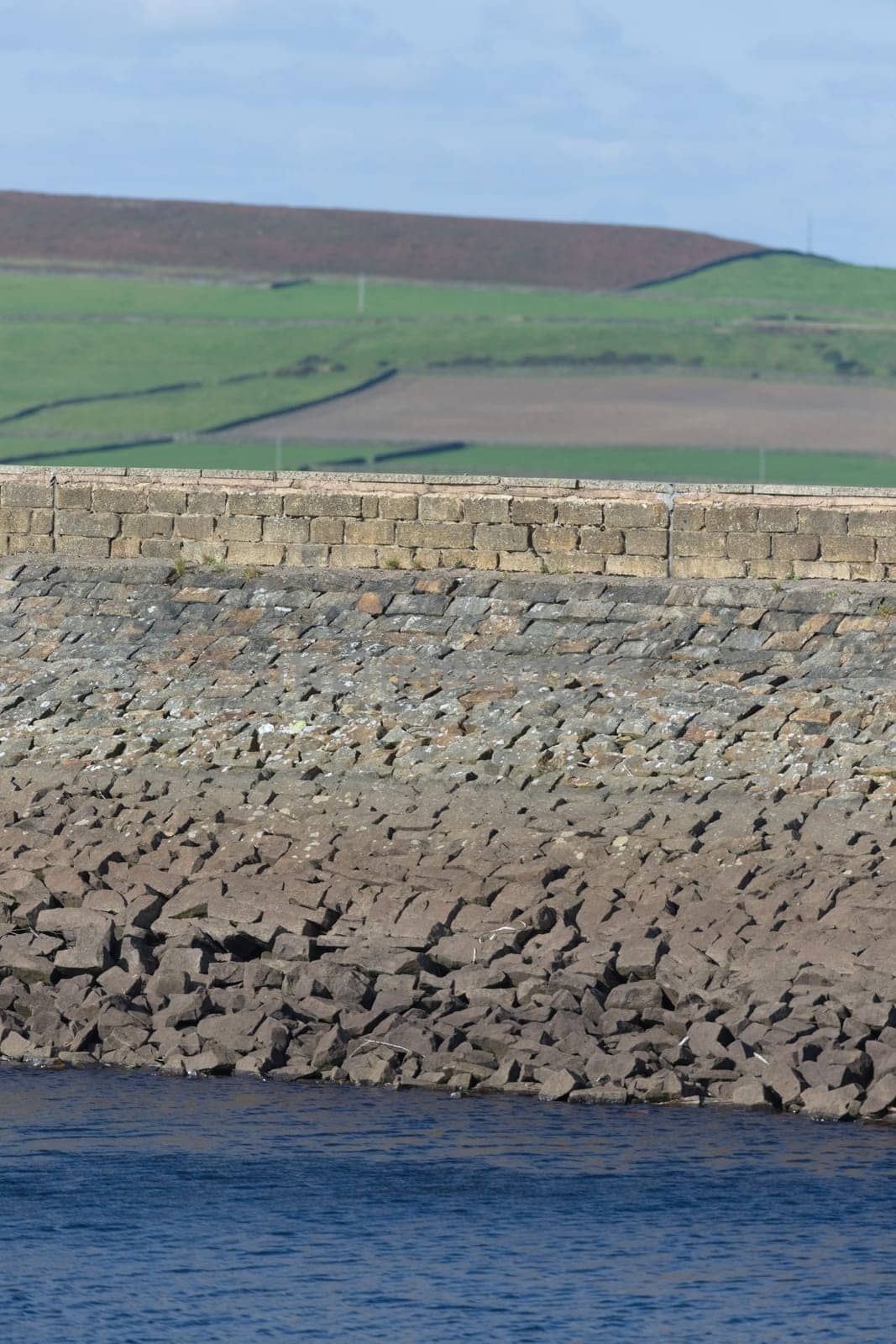 Vertical image of stone dam reservoir wall. Deep blue water and rural agricultural background are selectively out of focus. Warm sunny summers day