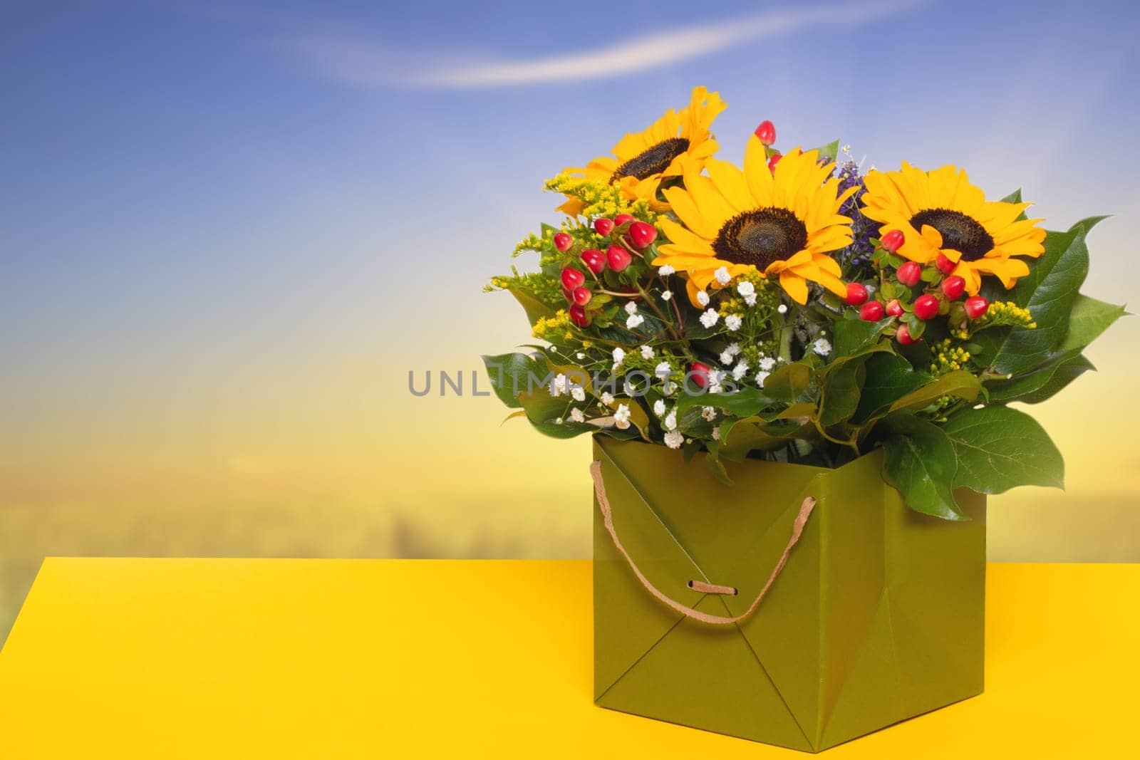 Closeup of a beautiful bouquet with yellow sunflowers in a decorative green gift box on a yellow table over a blurred blue yellow background. Macro. Space for design. Card concept.