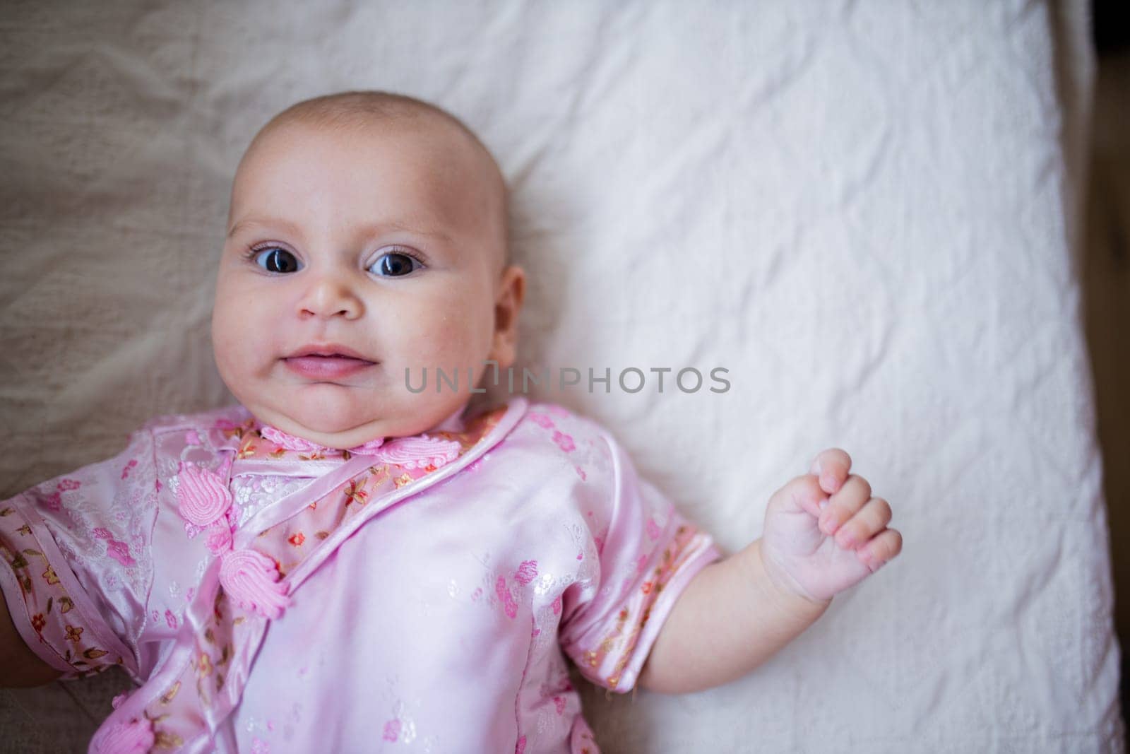 Curious adorable baby girl from above in Asian pink attire lying down on white bed. Portrait of cute female baby resting on bed. Happy babies on cradles and beds