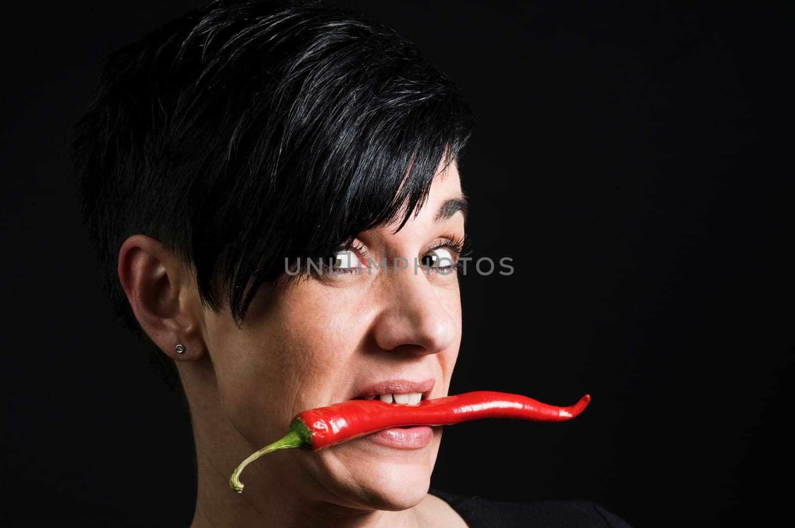 Lateral head portrait of a young black-haired woman in front of a black background holding a long red chilli pod across her teeth and looking skeptically into the camera.