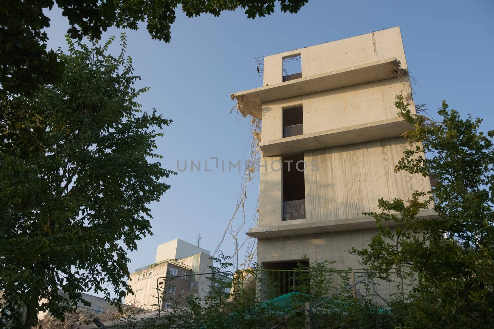 Steel cables and wires emerge from a partly demolished concrete building shell. Gentle morning sun and plain blue sky background. Leafy plant border.