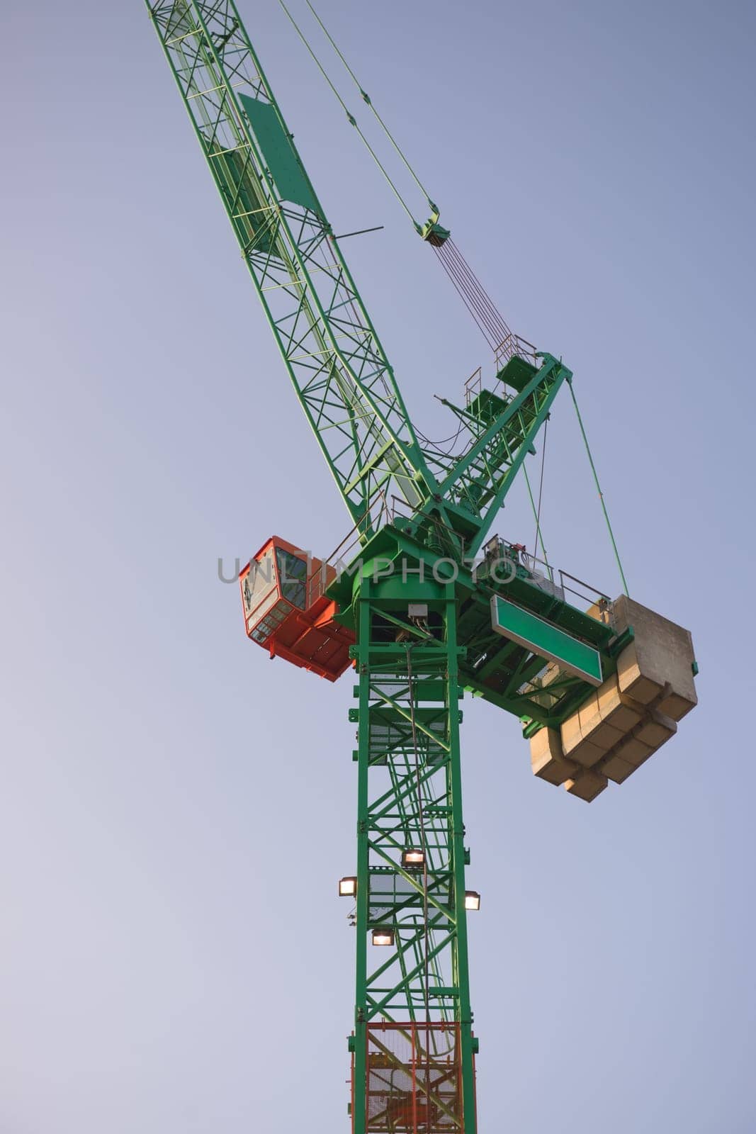 Vertical sunrise image looking up at a tall green and orange tower crane. Full frame crane and cabin with a plain blue sky background.
