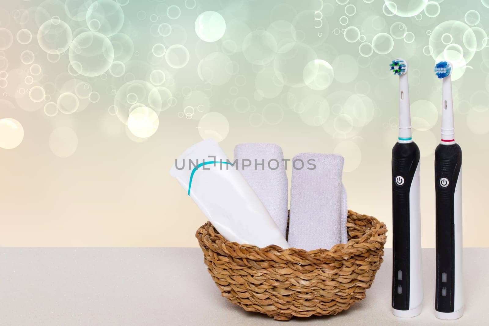 Two electrical tooth brushes, an oral care product or toothpaste and towels in a basket on a table against abstract blurred bright water bubbles background. Dental and healthcare concept. Copy space.