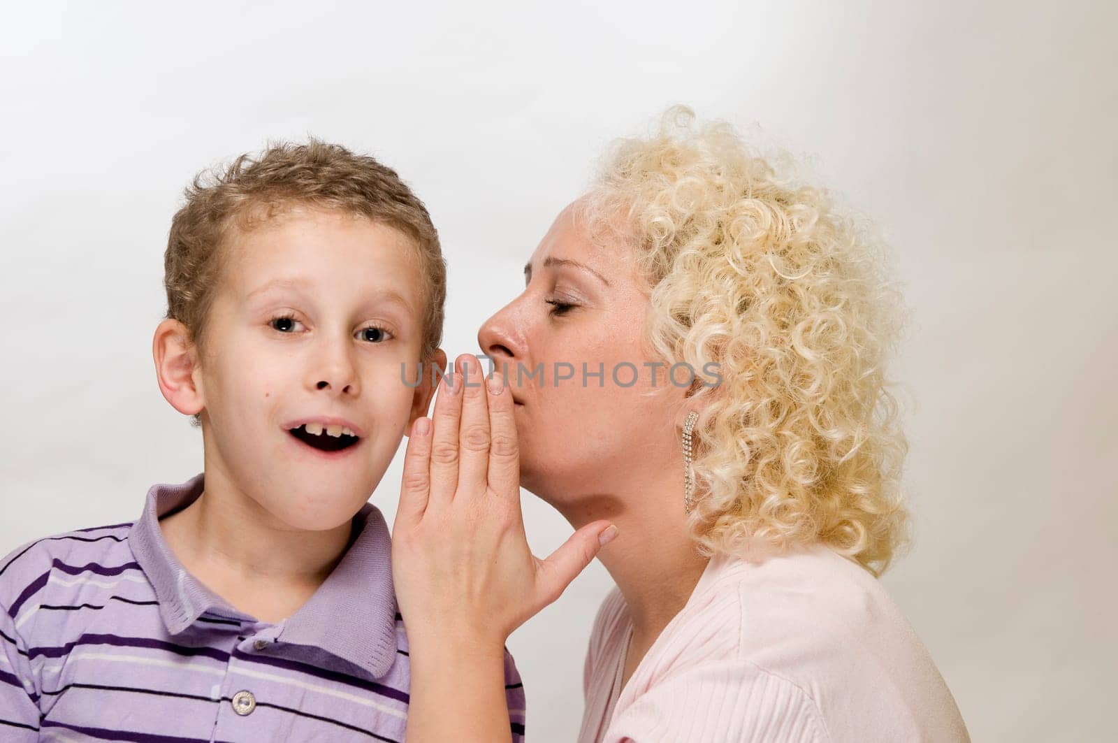 Head-and-shoulder view blonde mother whispers sideways into her 7-year-old son's ear with her hand held against a white background