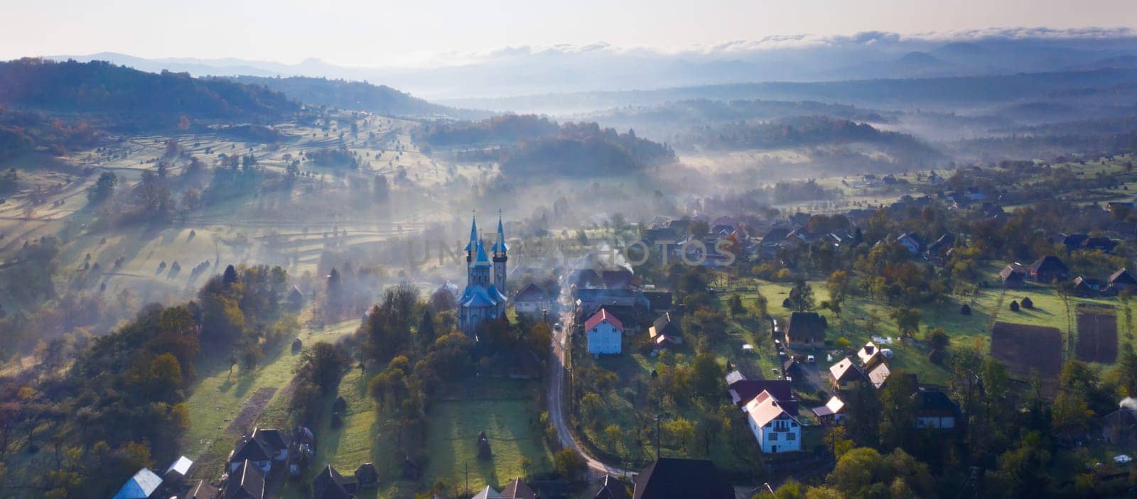 Aerial view of village in fog with golden sunbeams at sunrise in autumn. Beautiful rural landscape with foggy colorful trees, church, orange sky with sun. Fall in Romania.