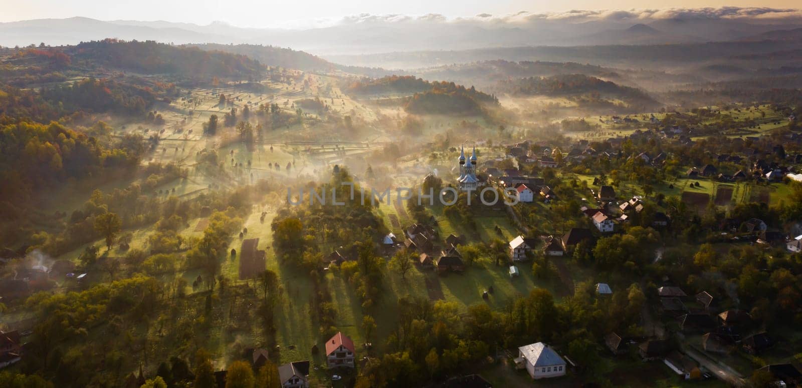 Aerial view of village in fog with golden sunbeams at sunrise in autumn. Beautiful rural landscape with foggy colorful trees, church, orange sky with sun. Fall in Romania.