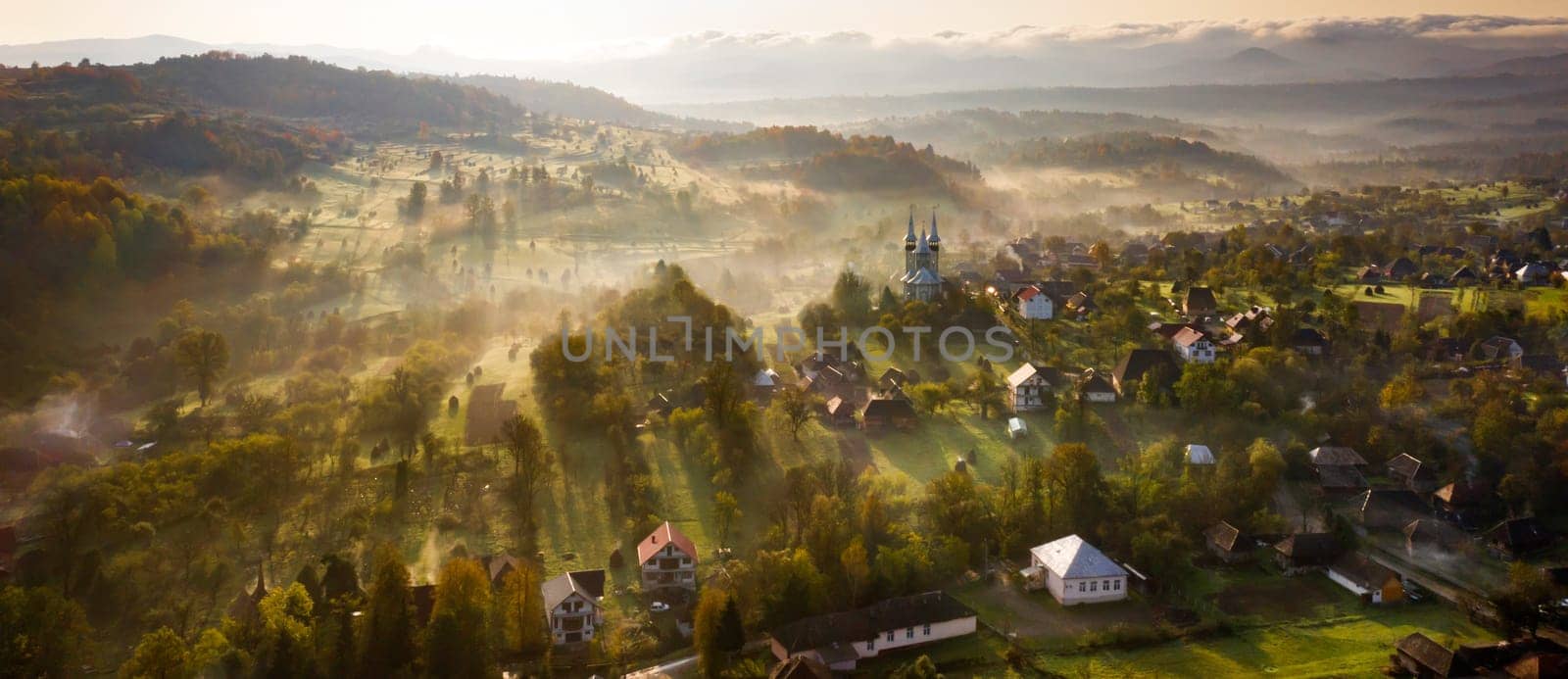 Aerial view of village in fog with golden sunbeams at sunrise in autumn. Beautiful rural landscape with foggy colorful trees, church, orange sky with sun. Fall in Romania.