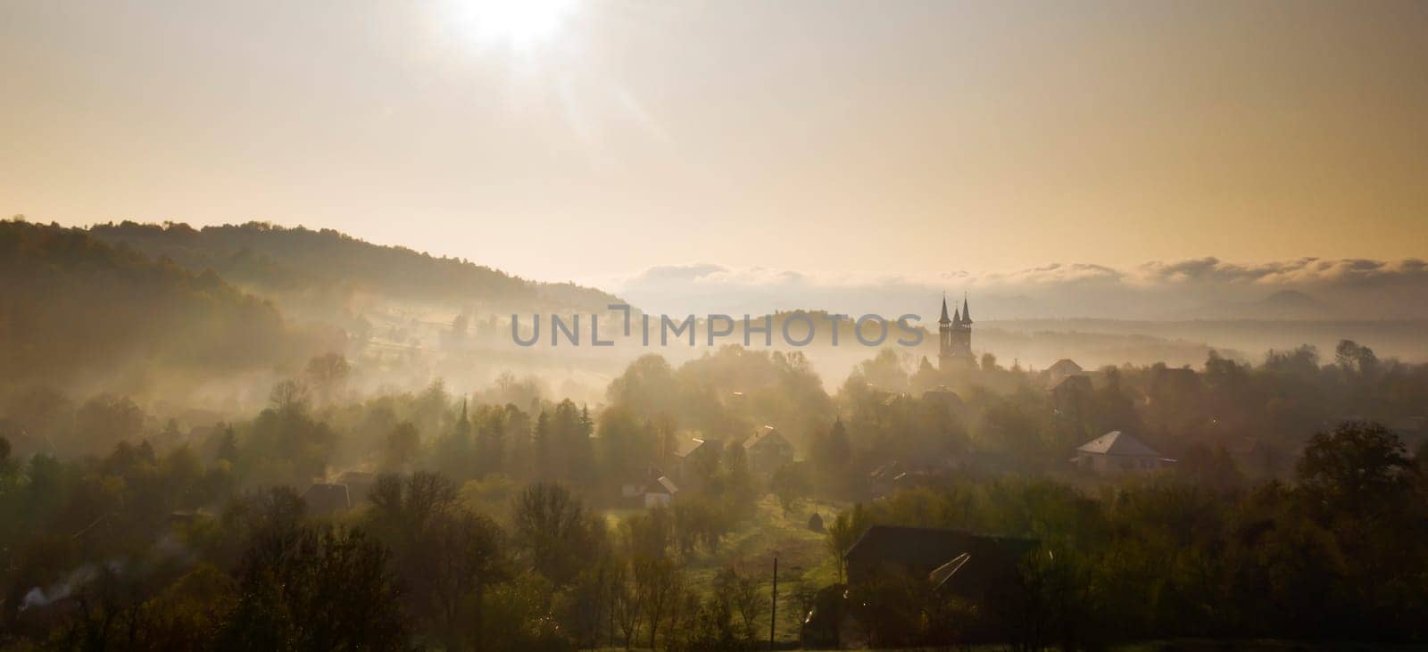 Aerial view of village in fog with golden sunbeams at sunrise in autumn. Beautiful rural landscape with foggy colorful trees, church, orange sky with sun. Fall in Romania.