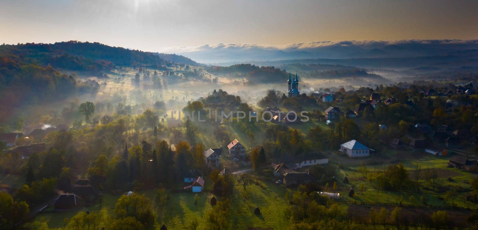 Aerial view of village in fog with golden sunbeams at sunrise in autumn. Beautiful rural landscape with foggy colorful trees, church, orange sky with sun. Fall in Romania.
