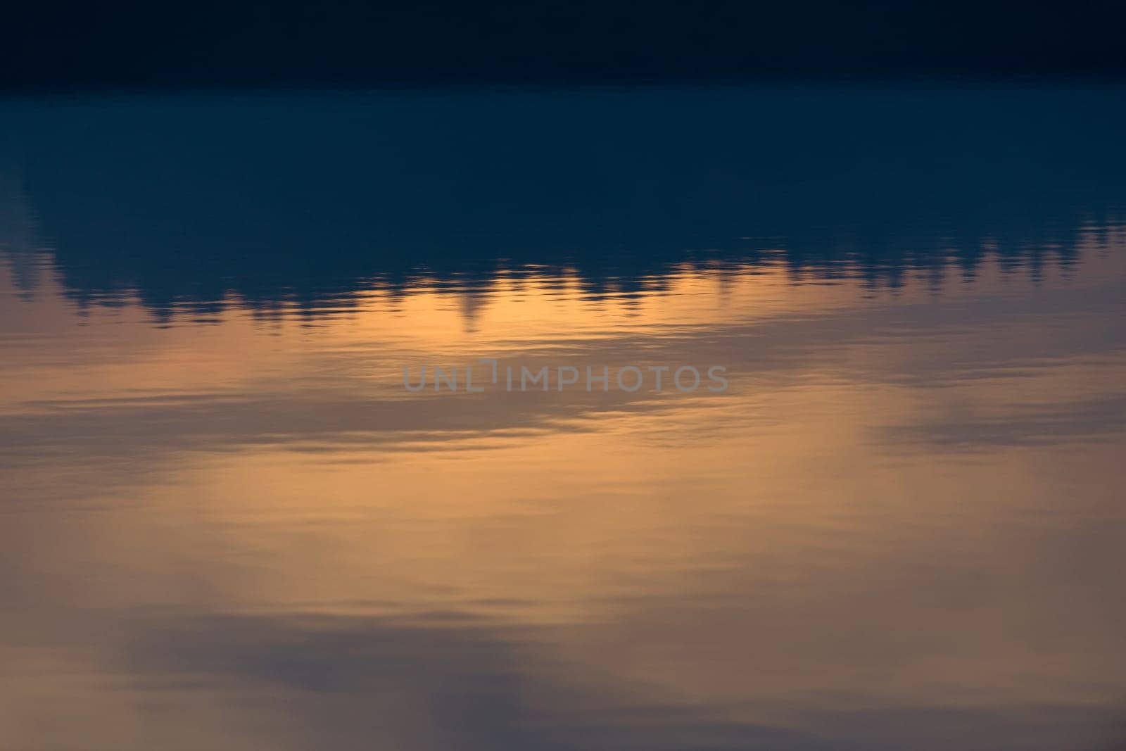 A warm orange glowing sunset behind a line of conifer trees is reflected and displayed upside down in the calm ripples of Redmires reservoir.