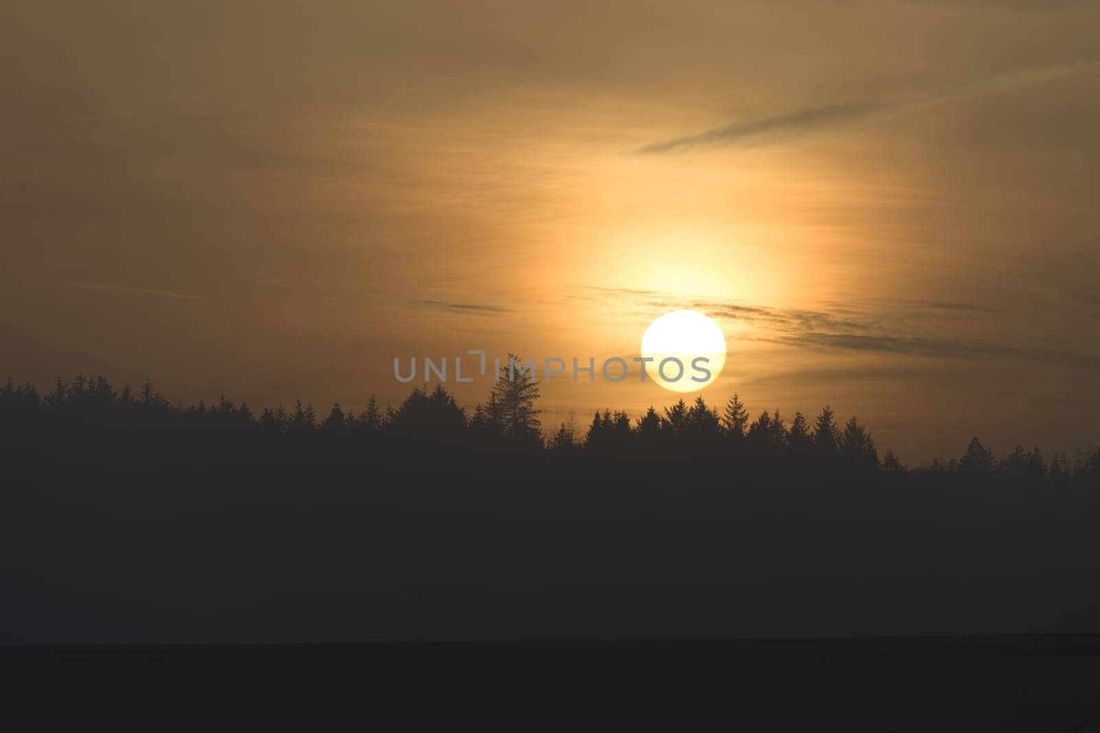 Telephoto sunset over a distant coniferous forest at redmires reservoirs near sheffield. Hazy sky as the sun just touches the tree tops. Copy space