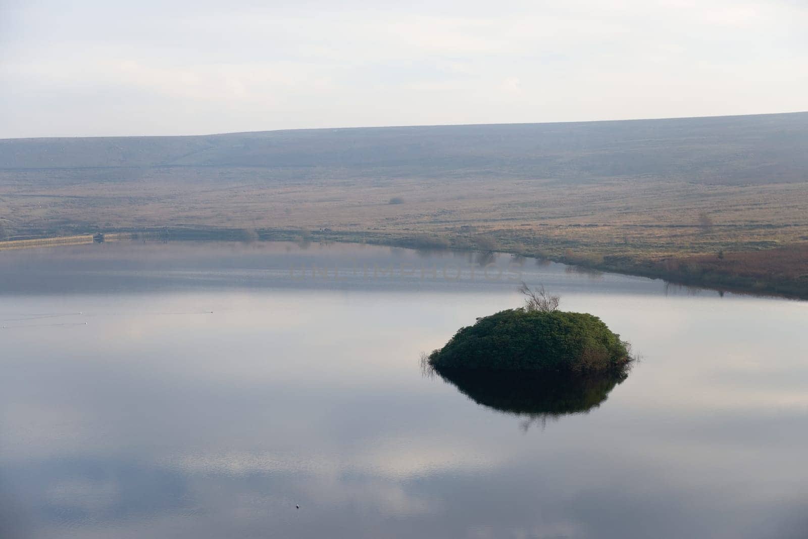 Middle reservoir of redmires reservoirs on a calm afternoon, reflecting a pale blue sky. Small overgrown island of plants stands out dark above water