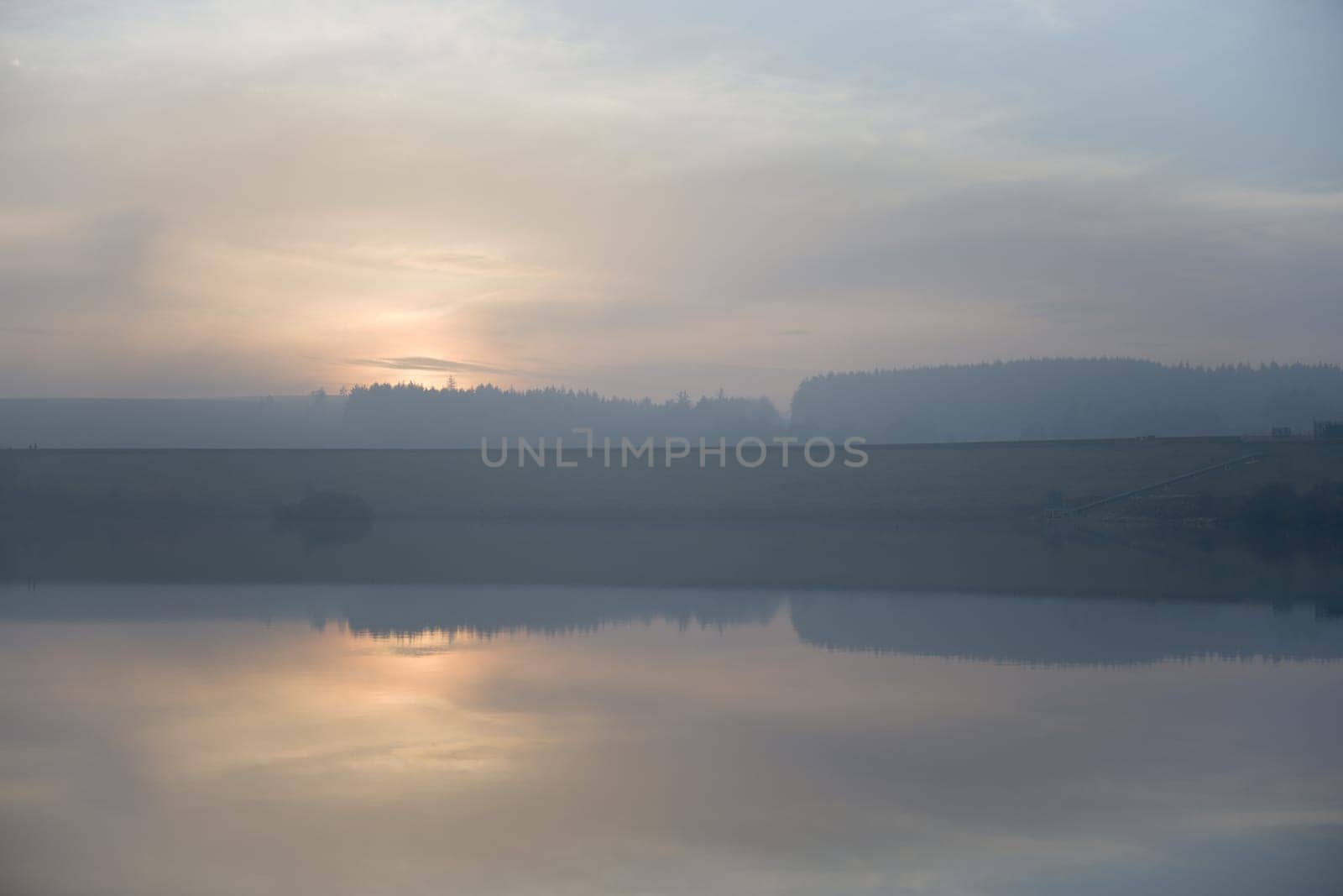 Calm reservoir water reflects misty sunset. Serene horizontal landscape image with blue and orange pastel colors. Hazy atmosphere with wispy clouds