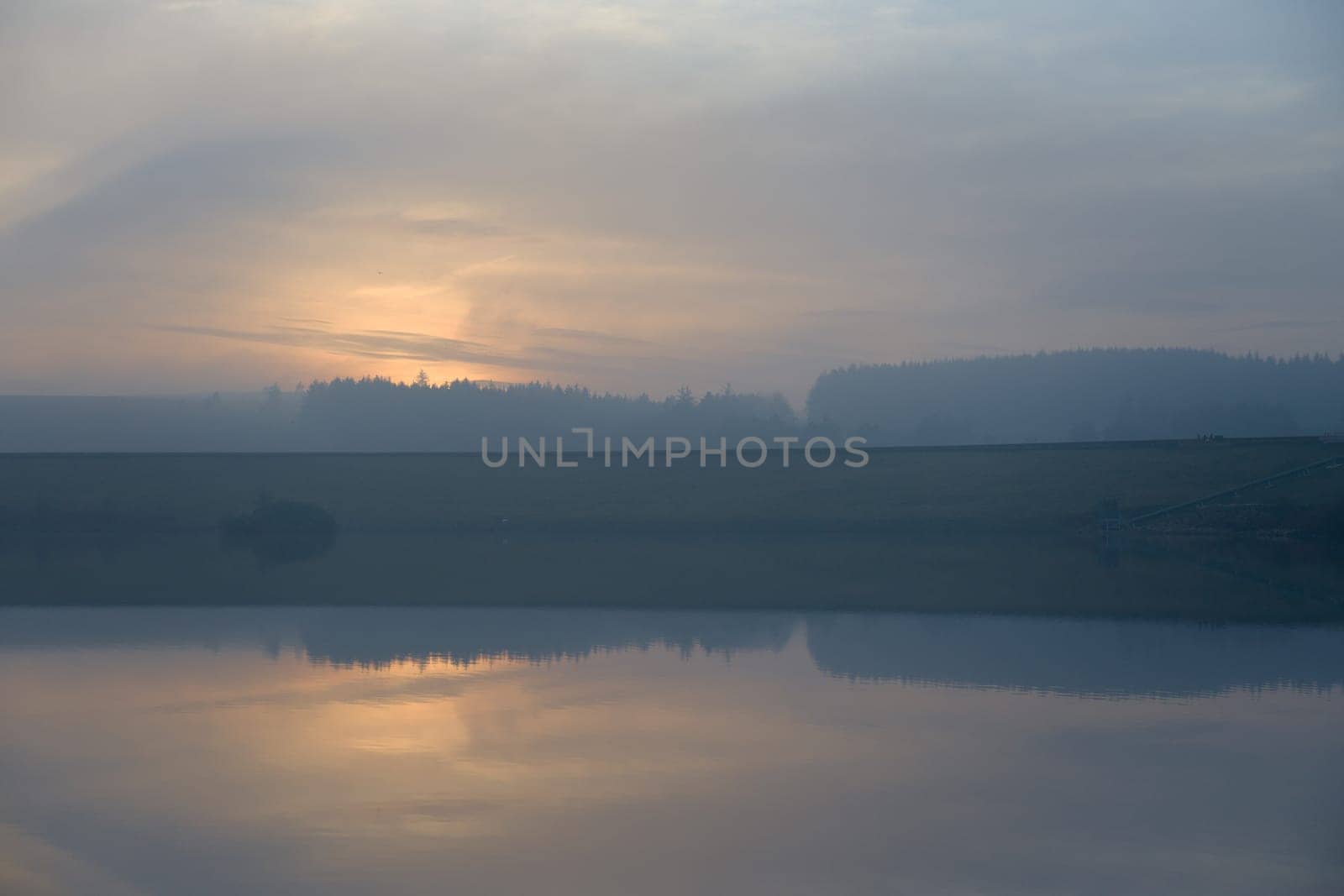 Gentle orange sunset silhouettes a coniferous forest. Dam wall divides the image in half, and the lower half of calm water reflects the sky above.