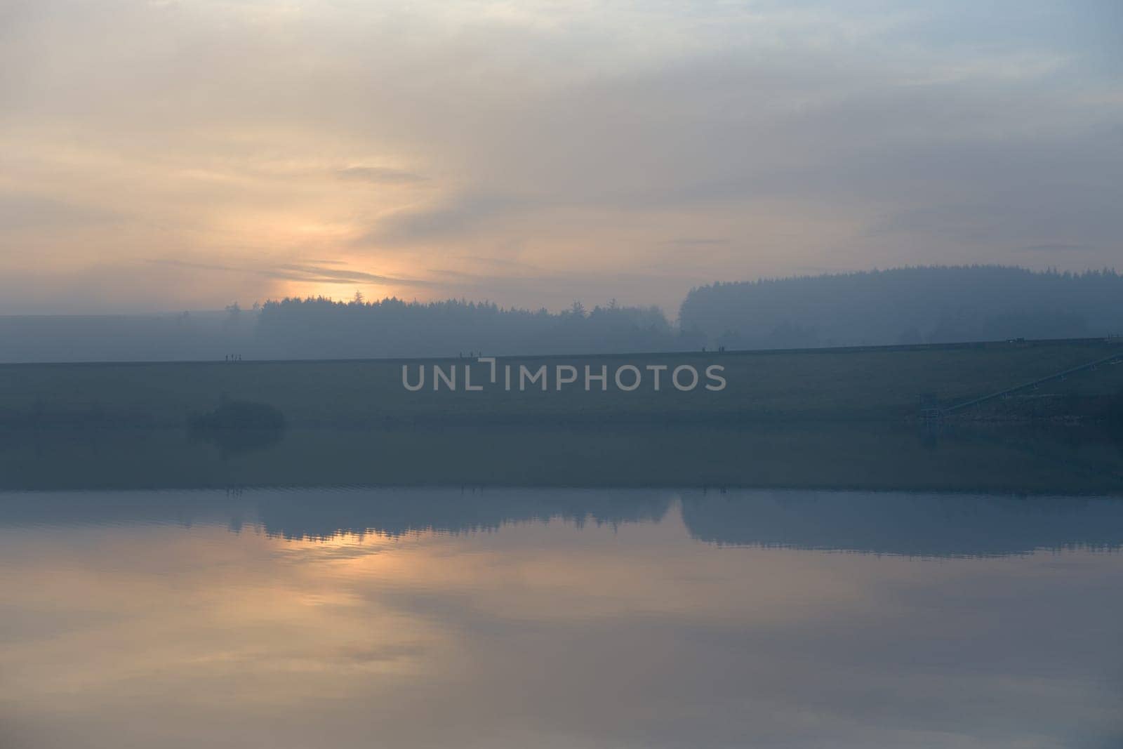 Golden sunset masked by haze and clouds is a back drop to 12 distant figures walking along redmires reservoirs dam wall. Cold day at the end of fall.