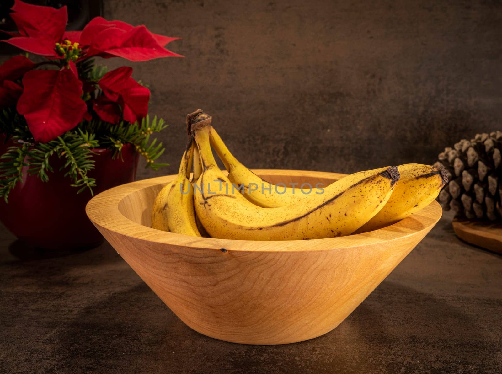 A turned rustic light wooden bowl with bananas stands on a dark surface with flowers in the background