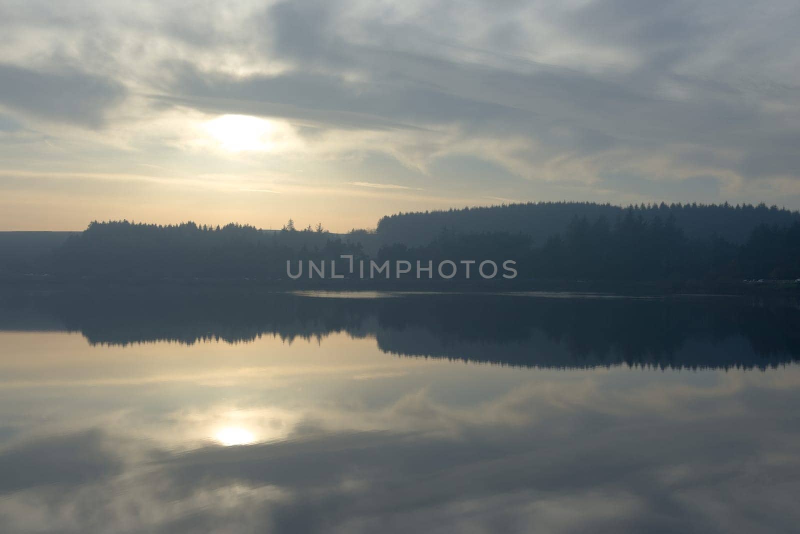 Winters sun and a cloudy sky reflected perfectly in the calm water of redmires reservoir. Hazy forest tree line in the distance, cars parked beneath