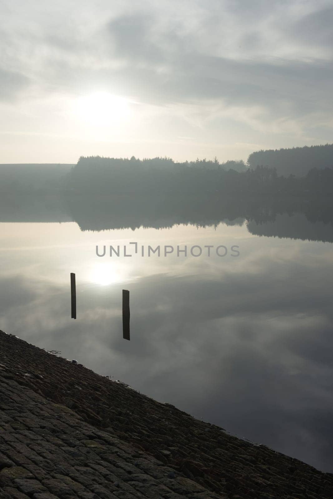 Two posts frame the winters sun as it is reflected in the calm water of a south yorkshire reservoir. Forest background and dam wall foreground.