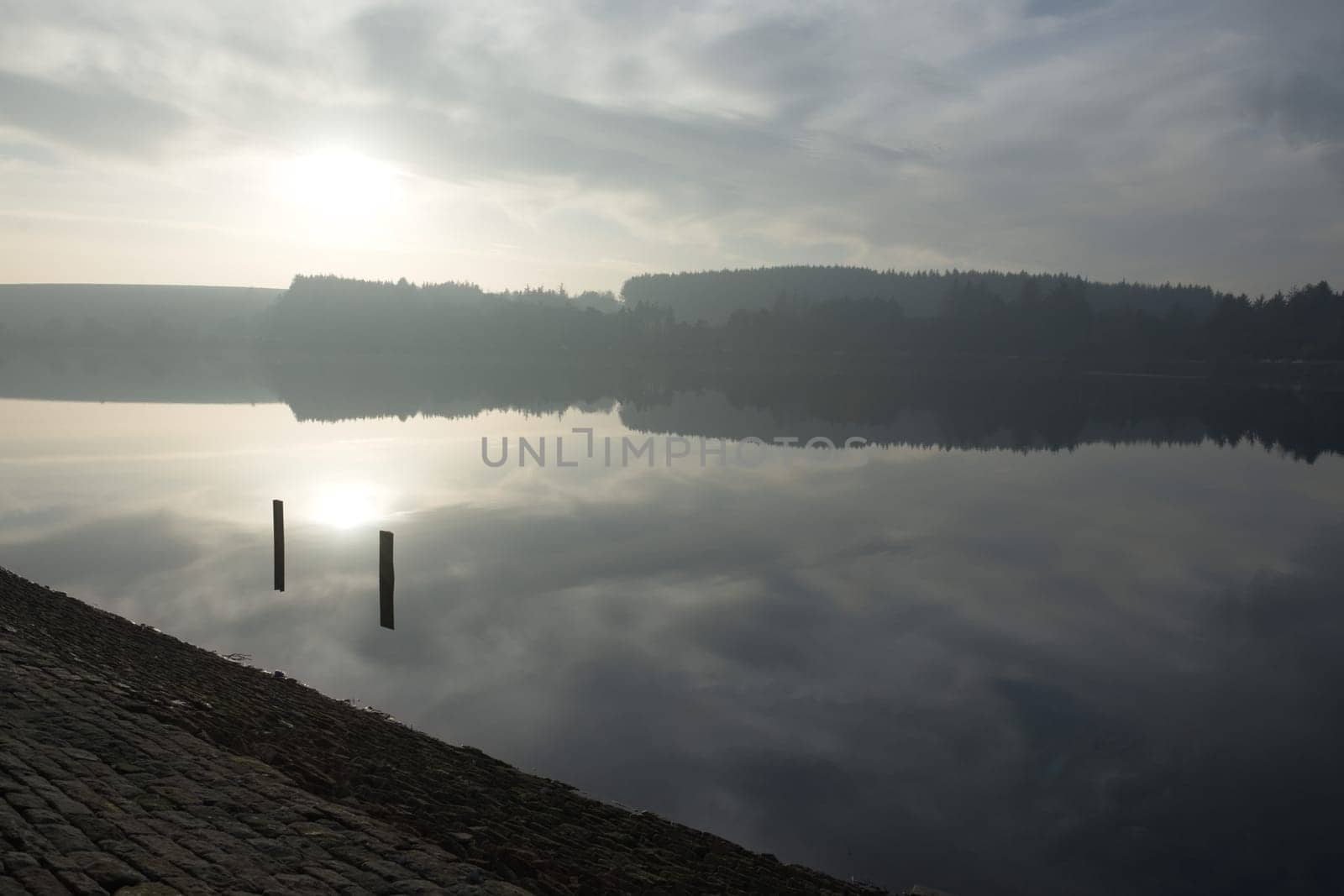 Large reservoir reflects the sky at sunset during winter. A distant forest of conifers is also mirrored in the shimmering water. Tranquil setting