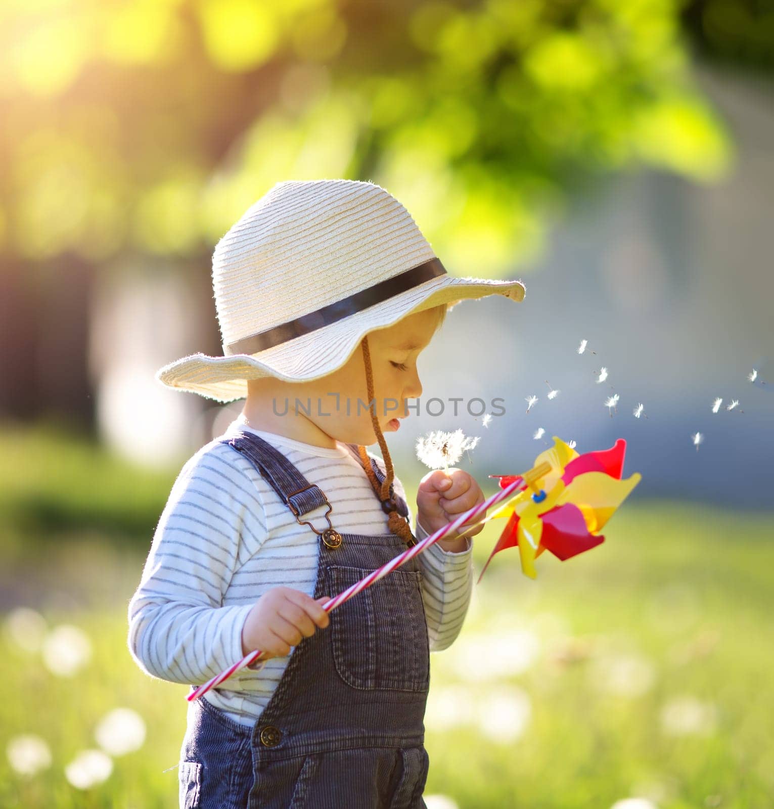 Happy baby boy standing in grass on the fieald with dandelions at sunny summer evening. Smiling child outdoors
