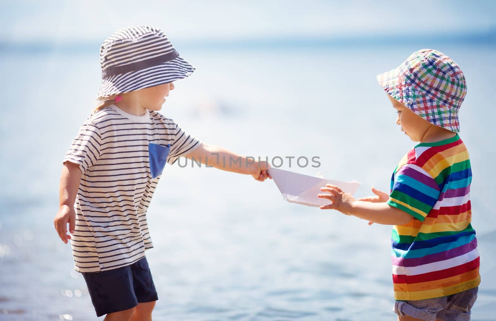 Girl and boy playing on the beach in summer hats and holding paper ships. Happy children running at seaside