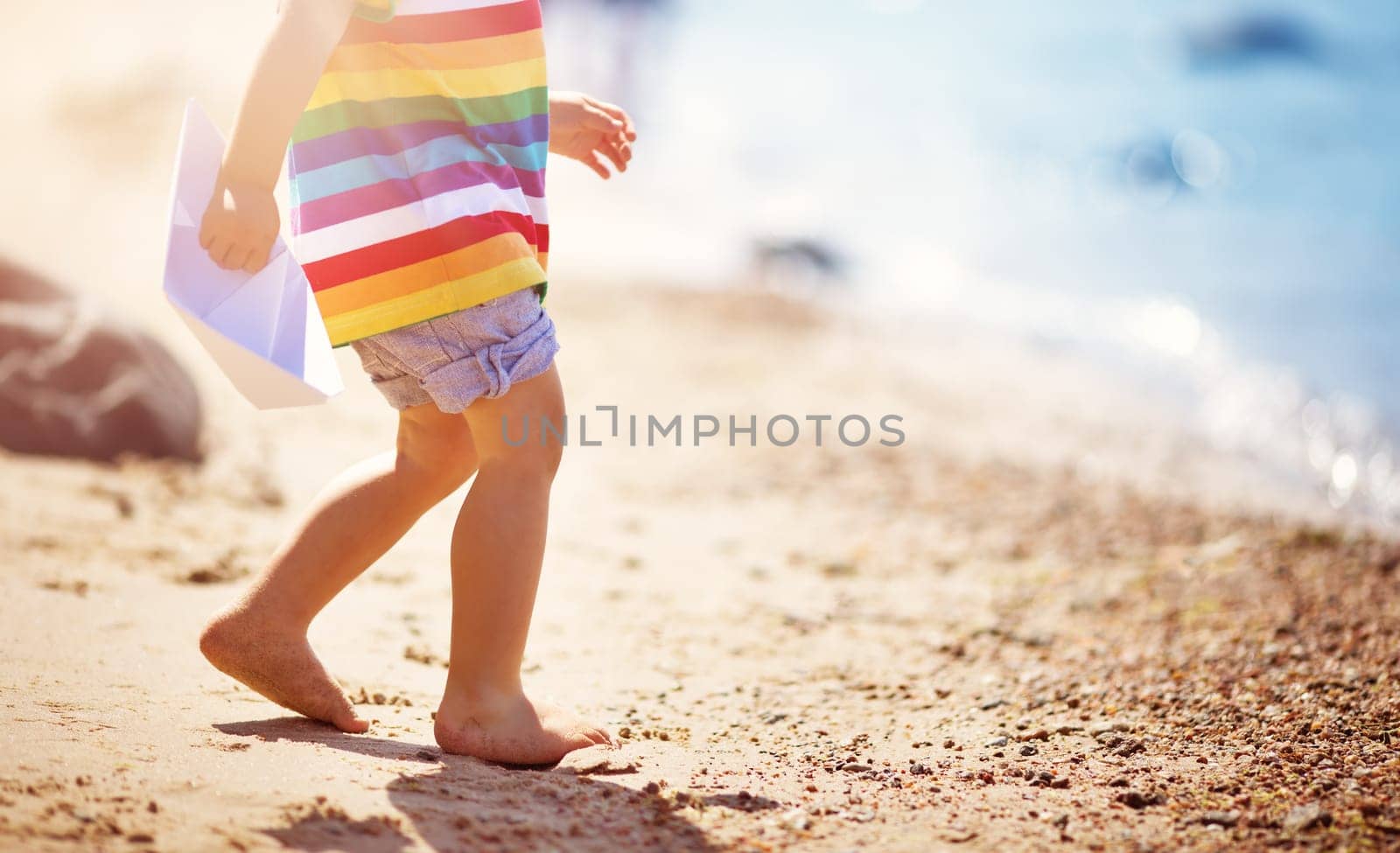 two years old boy playing at the beach in hat. Child with a paper ship at sea