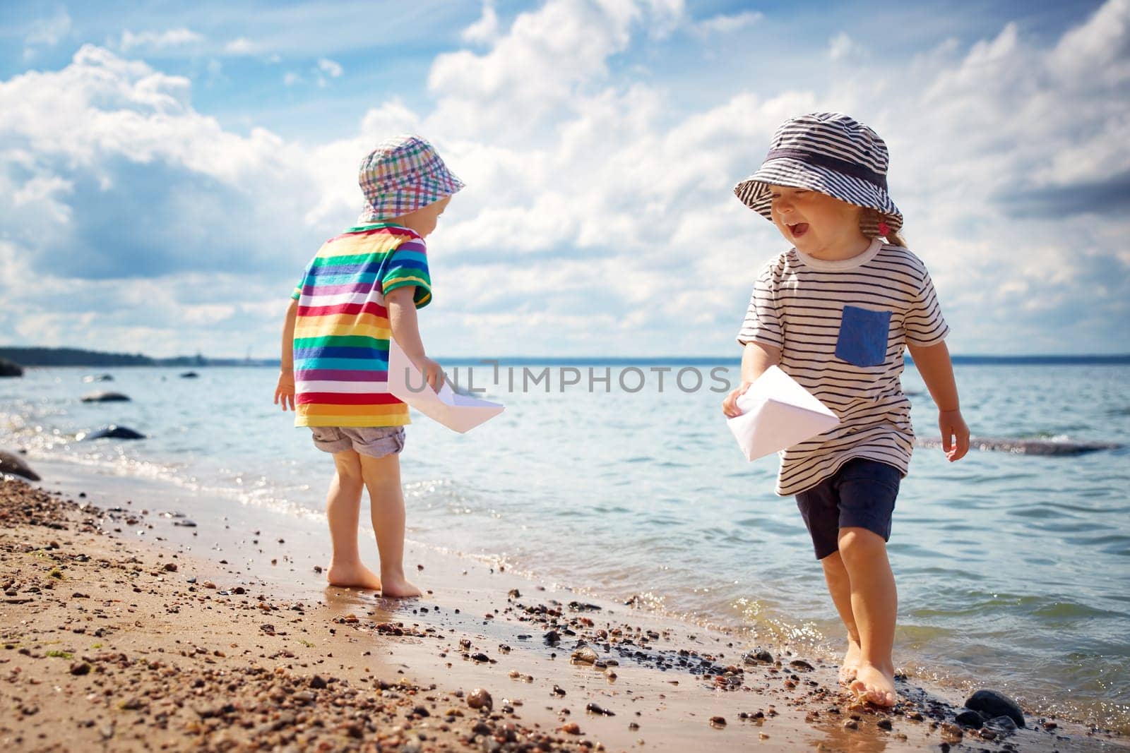 Girl and boy playing on the beach in summer hats and holding paper ships. Happy children running at seaside
