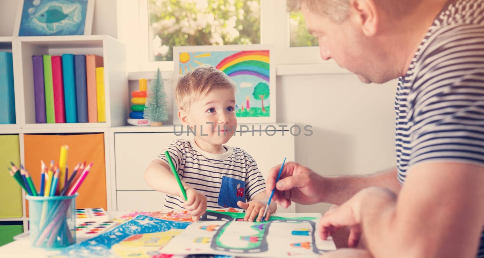 Little child drawing on the paper with father. Boy sitting on the chair at table with paints with dad