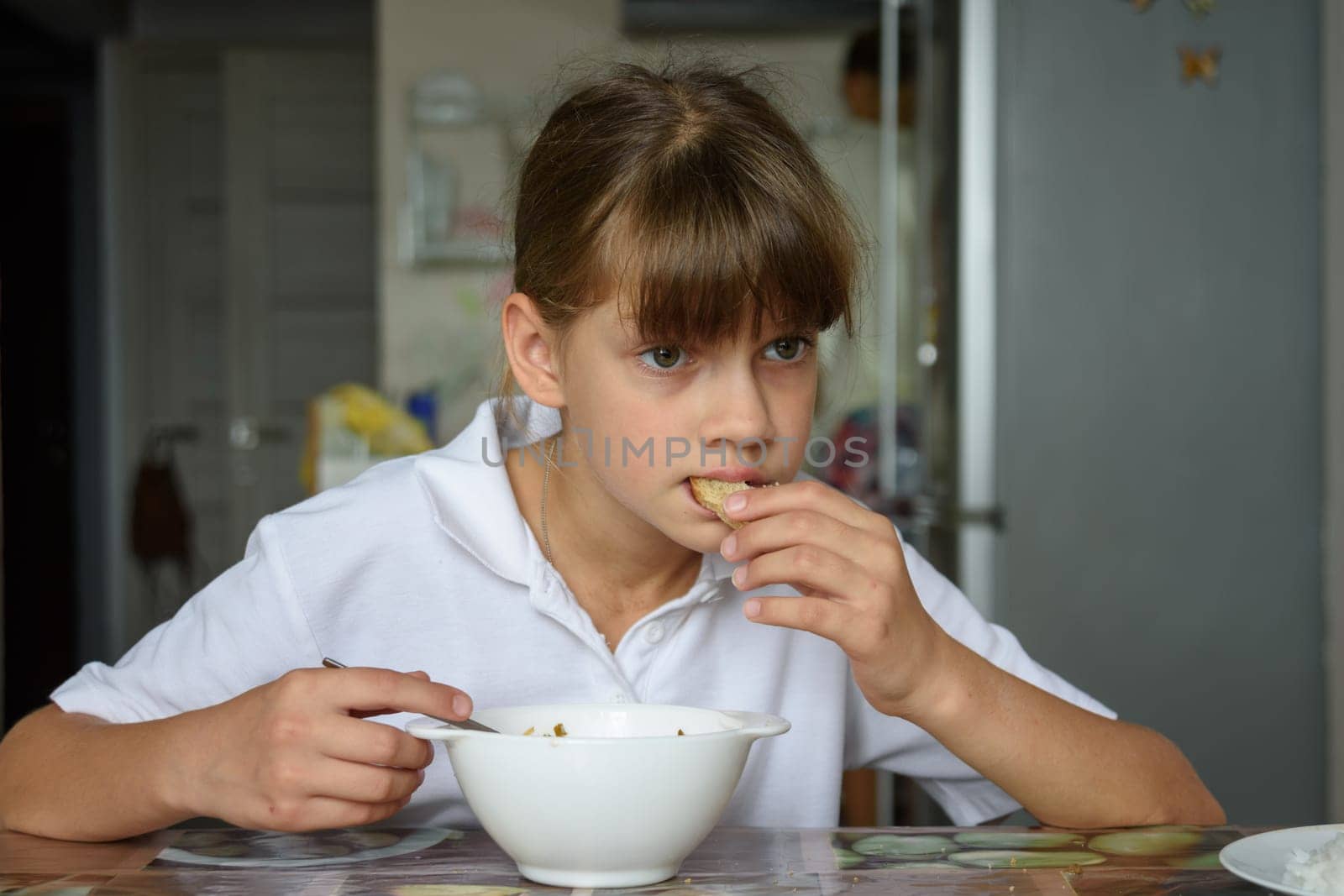 Tired hungry schoolgirl eating soup at the table in the kitchen