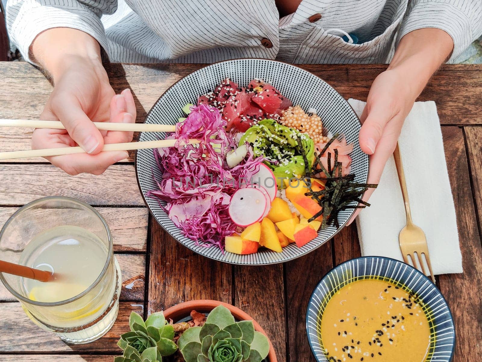Woman eating tasty colorful healthy natural organic vegetarian Hawaiian poke bowl using asian chopsticks on rustic wooden table. Healthy natural organic eating concept.