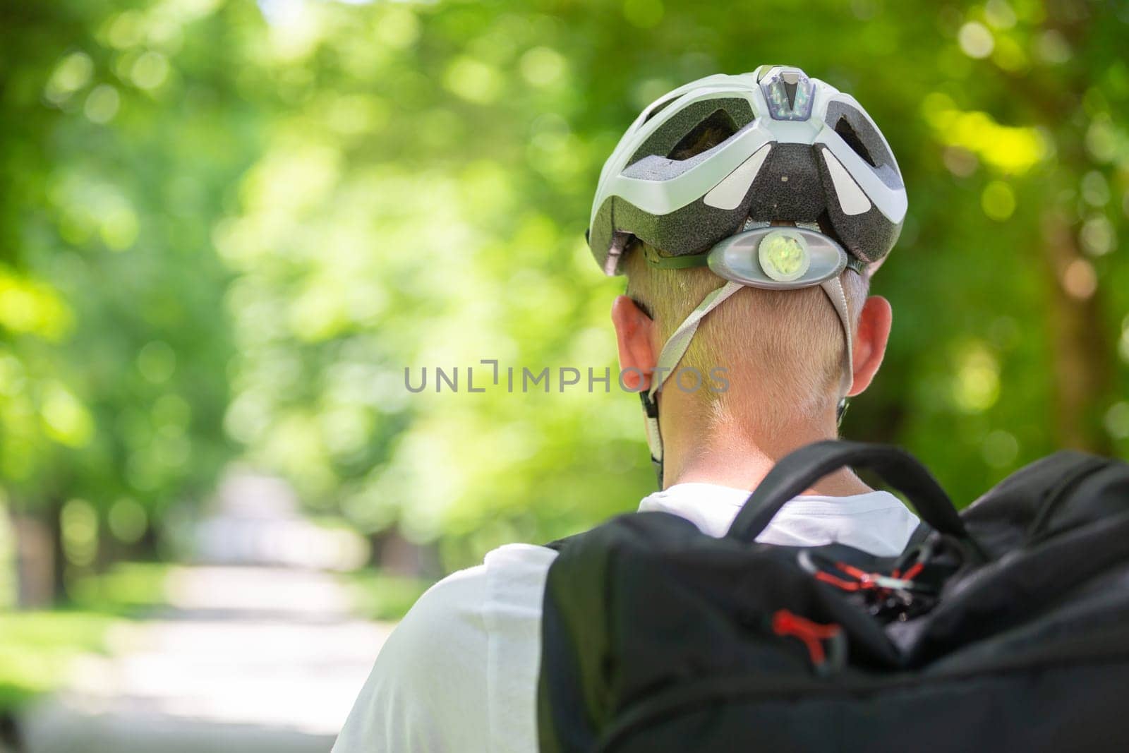 Rear view of unrecognizable man wearing protective helmet on bicycle cycling in city park.