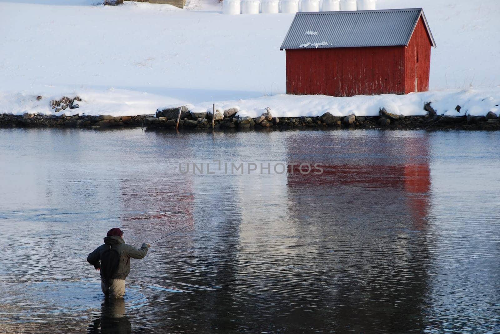 A fly fisherman in waders in Norwegian waters. In front of him the snow-covered area and a typical Norwegain red wodden house.