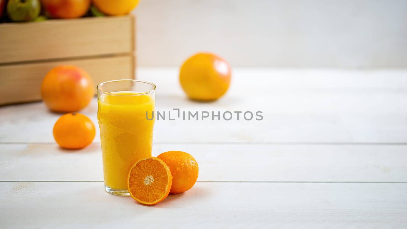 Glass of juice with cut orange on white desk with copy space. Cup of fresh yellow bevarage with grapefruits and wooden box in the background. Still life of sweet healthy drink with fruit with space for text.