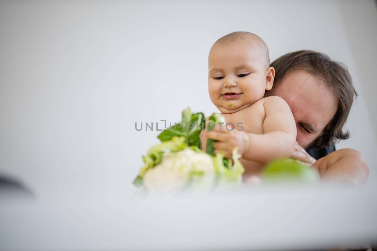 Father lovingly holding and kissing his happy baby daughter above table. Adorable baby smiling and playing with cauliflower. Babies interacting with food