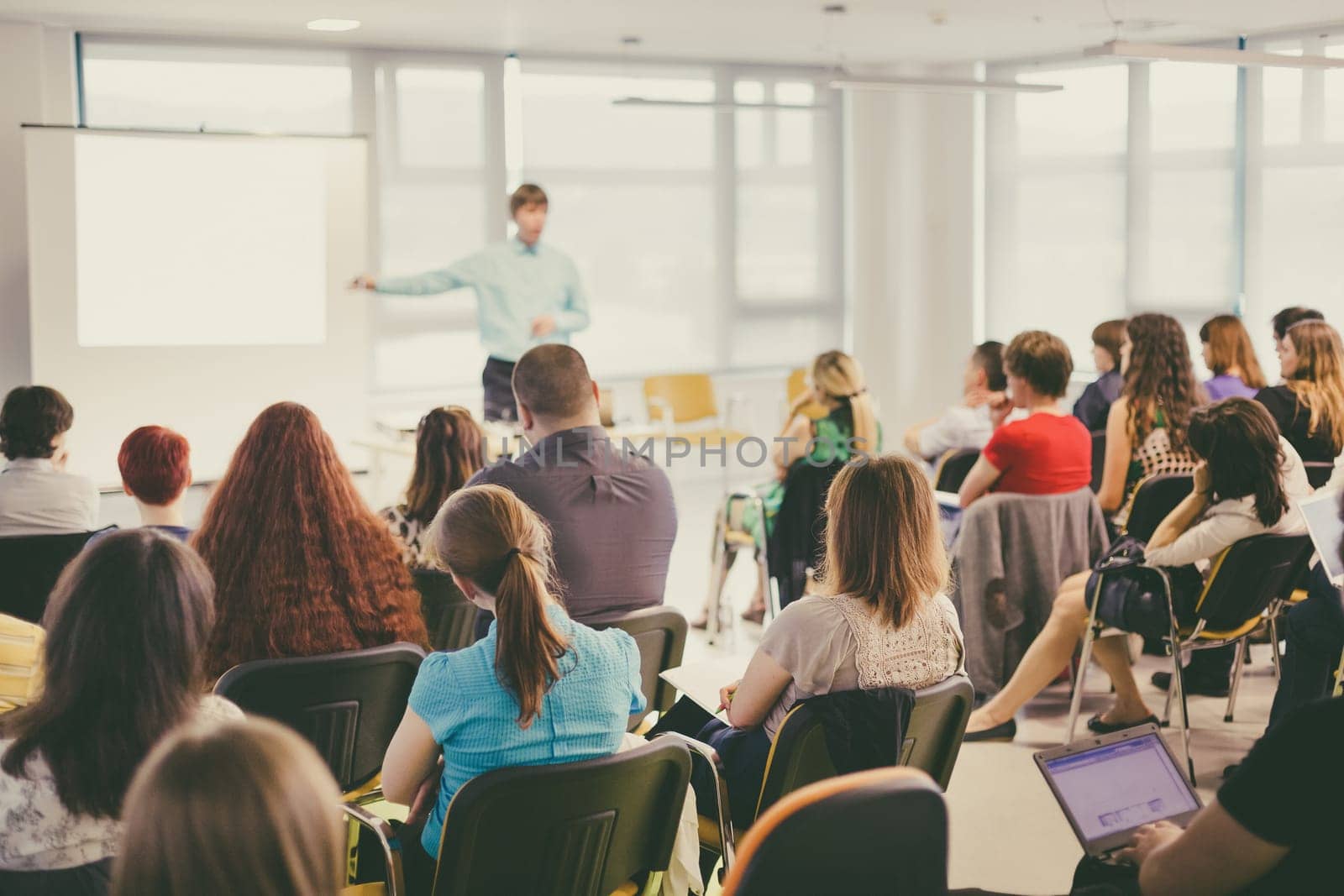 Business and entrepreneurship symposium. Speaker giving a talk at business meeting. Audience in conference hall. Rear view of unrecognized participant in audience.