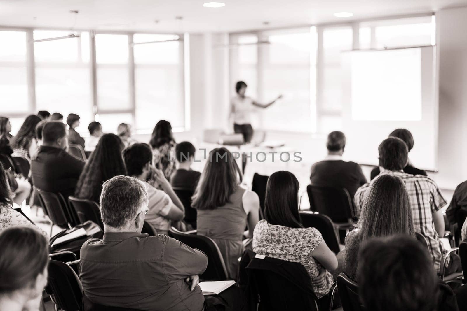 Business and entrepreneurship symposium. Speaker giving a talk at business meeting. Audience in conference hall. Rear view of unrecognized participant in audience.