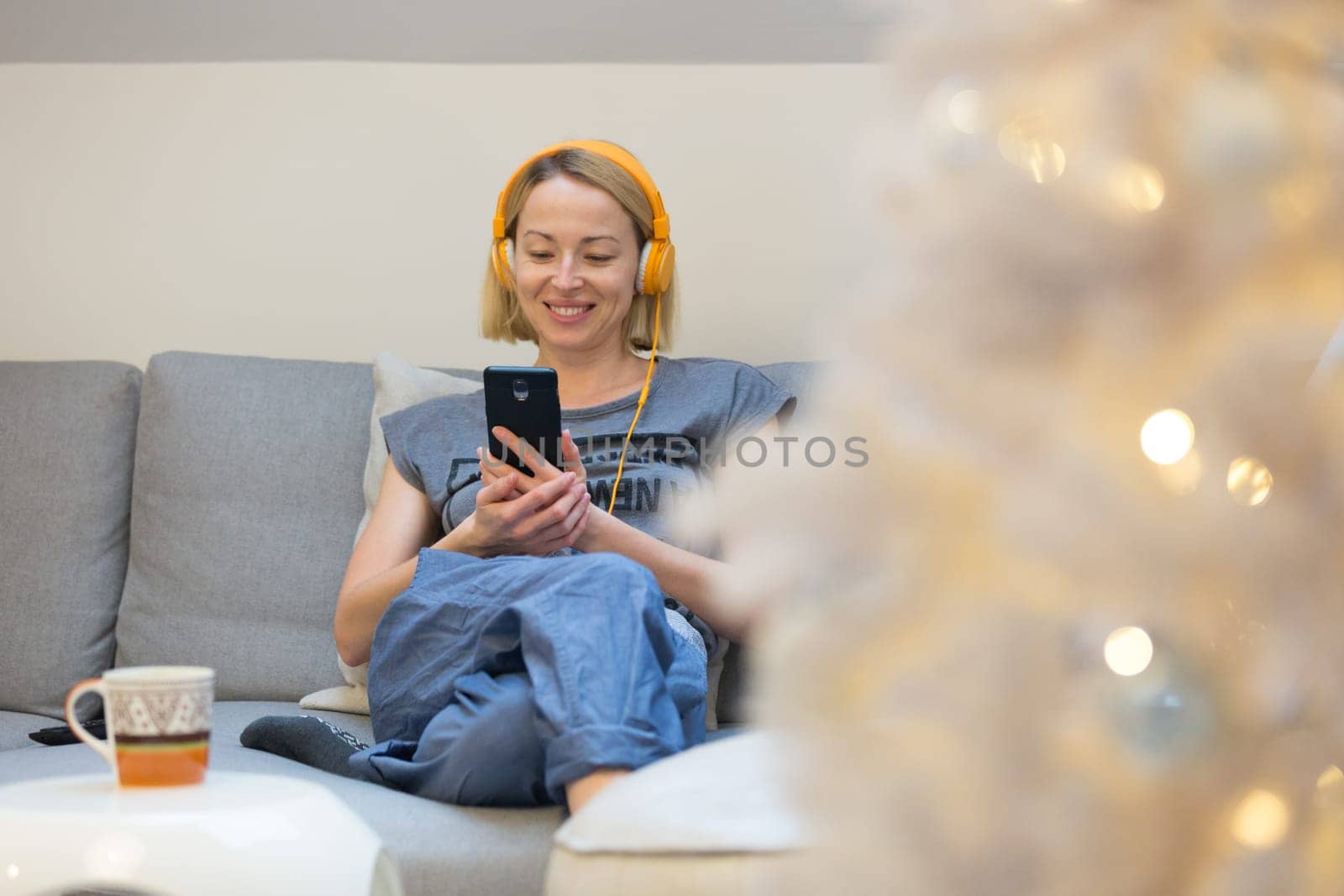 Young cheerful woman sitting indoors at home living room sofa using social media on phone for video chatting and staying connected with her loved ones. Stay at home, social distancing lifestyle.