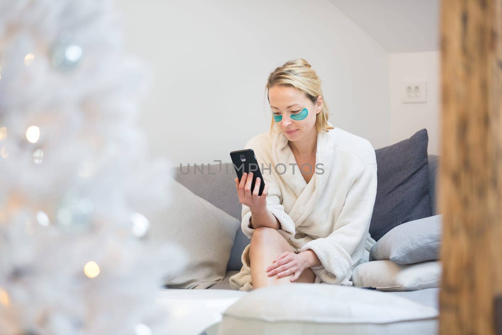 Woman wearing cosy warm bathrobe and cosmetic eye patches relaxing on sofa using social media on phone for video chatting with her loved ones in winter Christmas time.
