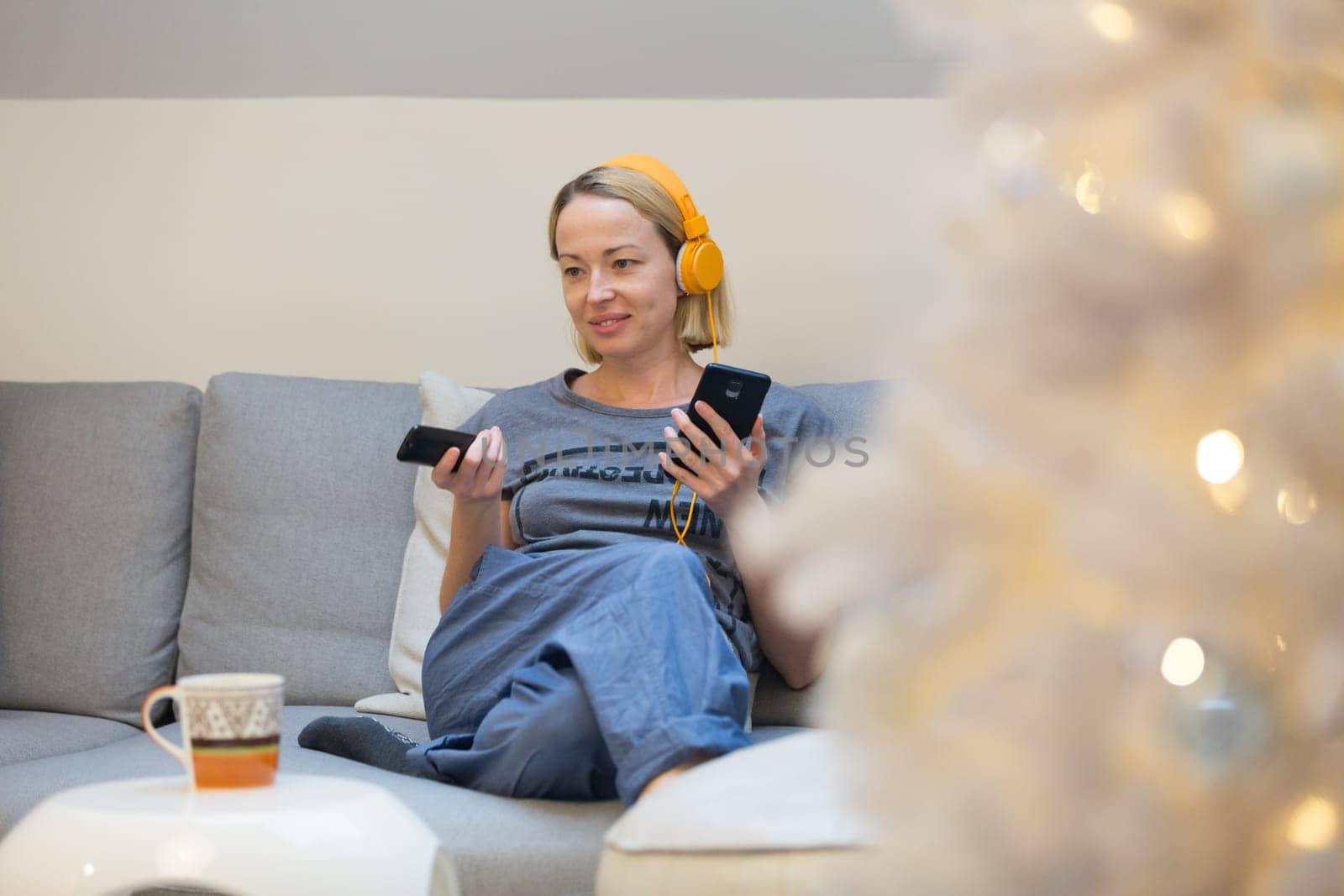 Young cheerful woman sitting indoors at home living room sofa watching TV, using social media on phone for video chatting and staying connected with her loved ones.
