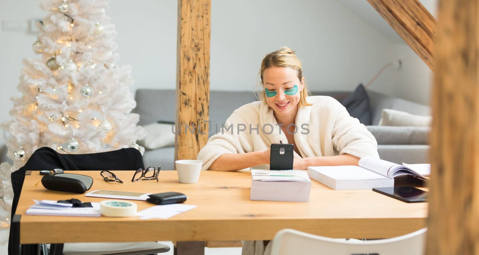 Young businesswoman wearing cosy warm bathrobe and cosmetic eye patches working remotly from home in winter Christmas time during 2020 corona virus pandemic. Work from home, selter in place, concept.