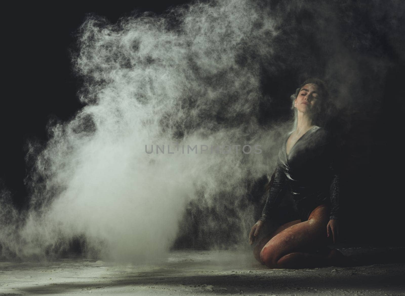 young beautiful caucasian woman with long hair is dressed in a sports black bodysuit and sits on the floor and throwing white flour up, black background