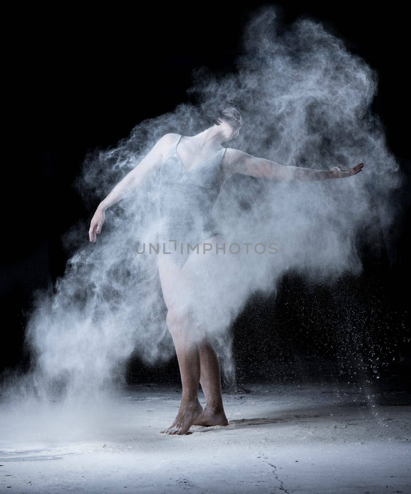 woman with a sports figure dancing in a cloud of white scattered flour on a black background, the dancer is dressed in a black bodysuit