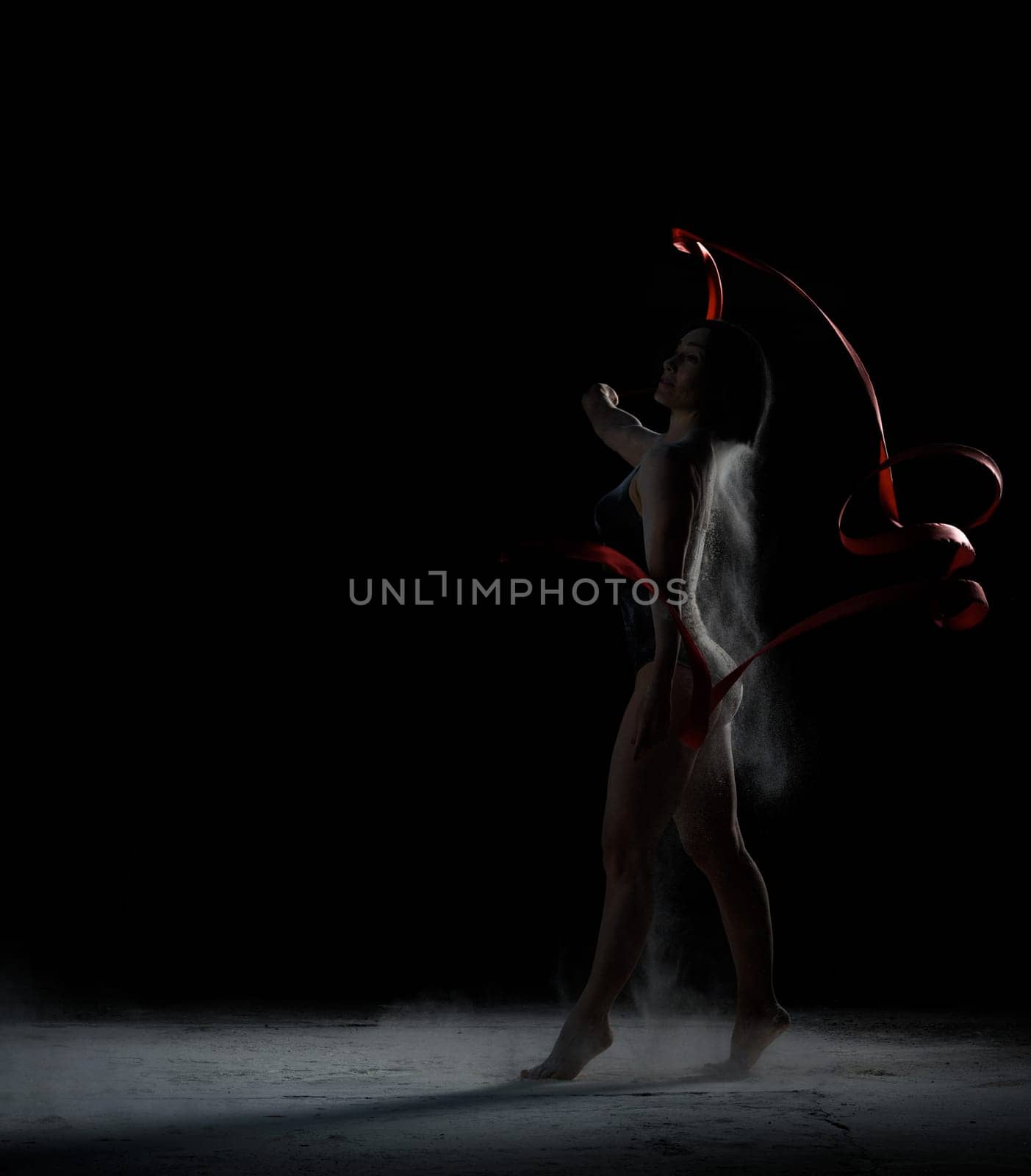 beautiful caucasian woman in a black bodysuit with a sports figure dancing with red ribbon on a black background, particles of flour fly in different directions