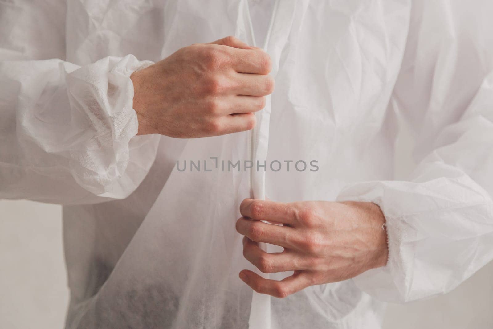 Doctor epidemiologist puts on white protective suit, fastens a zipper, hands close-up. Hands in protective rubber gloves. Personal protective equipment concept.