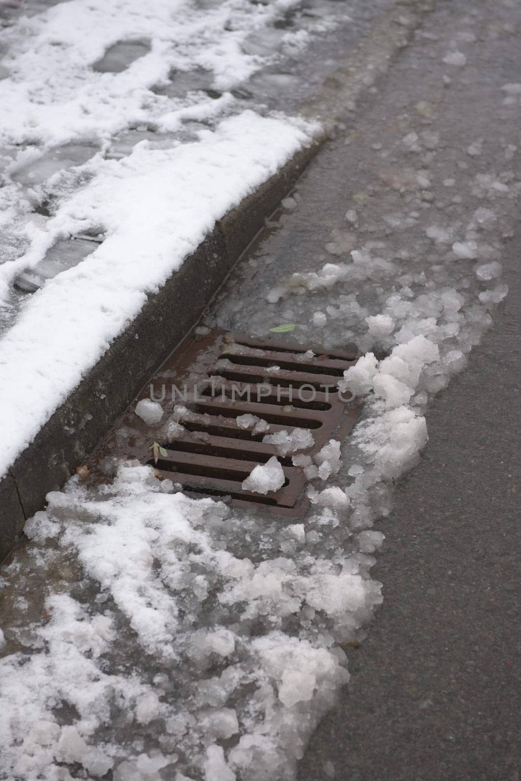 7 - Urban highway drain covered in thawing snow fall. Water runs along the curbside and into the surface storm drain. This area is at risk of flooding