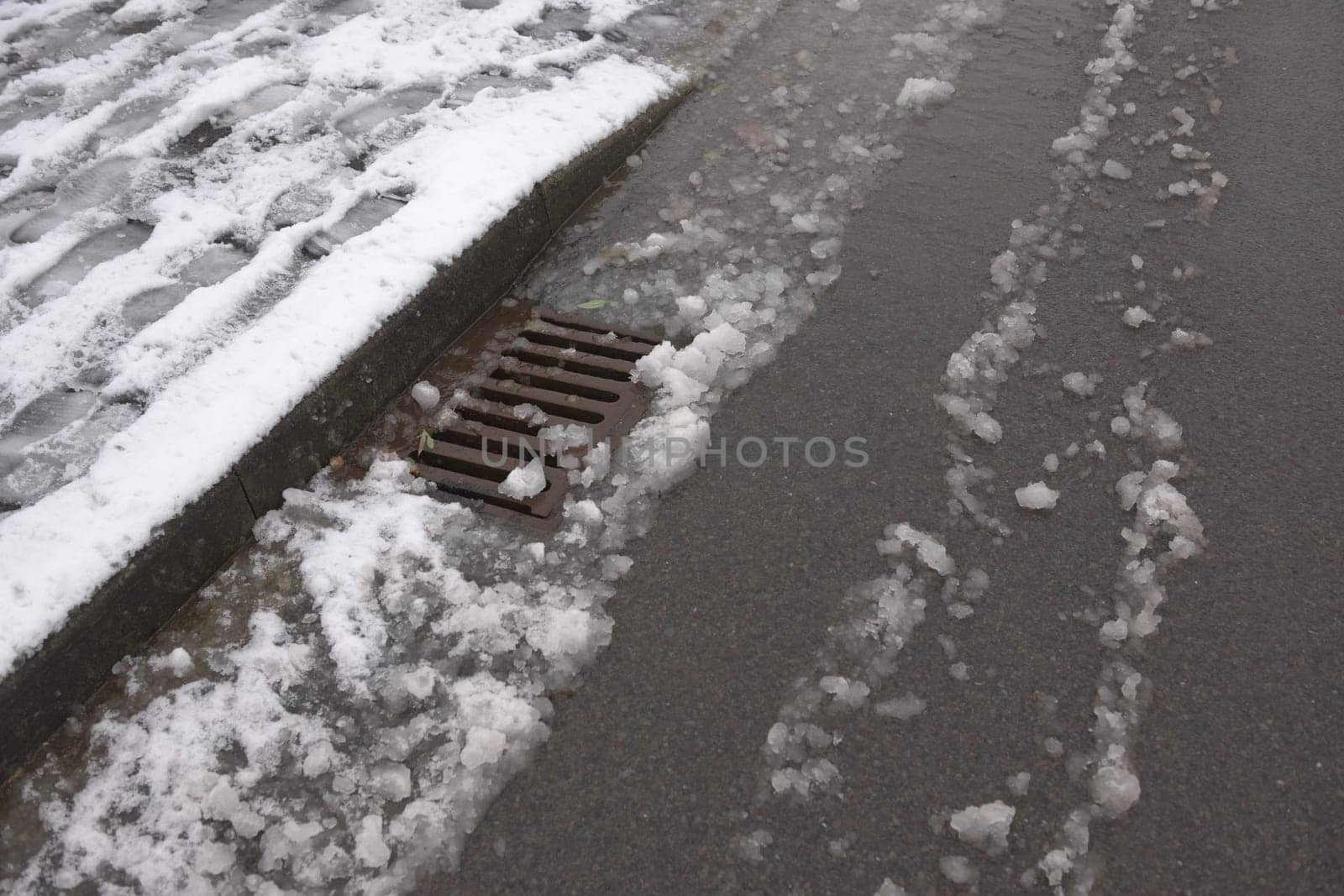 5 - Urban tarmac road and sidewalk covered in heavy snow that is beginning to thaw. Surface level storm drain deals with the resulting water.