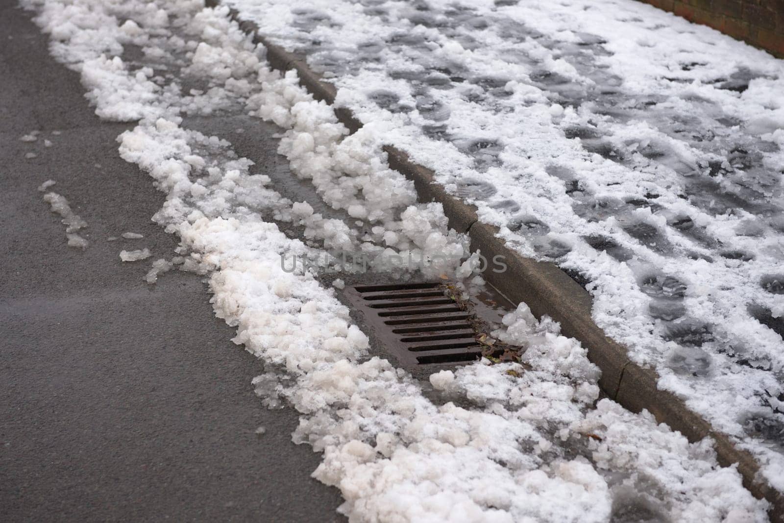 12 - Urban scene of footsteps in a pavement full of snow. Drain emerges from the accumulation of snow at the road side. Winter environmental scene