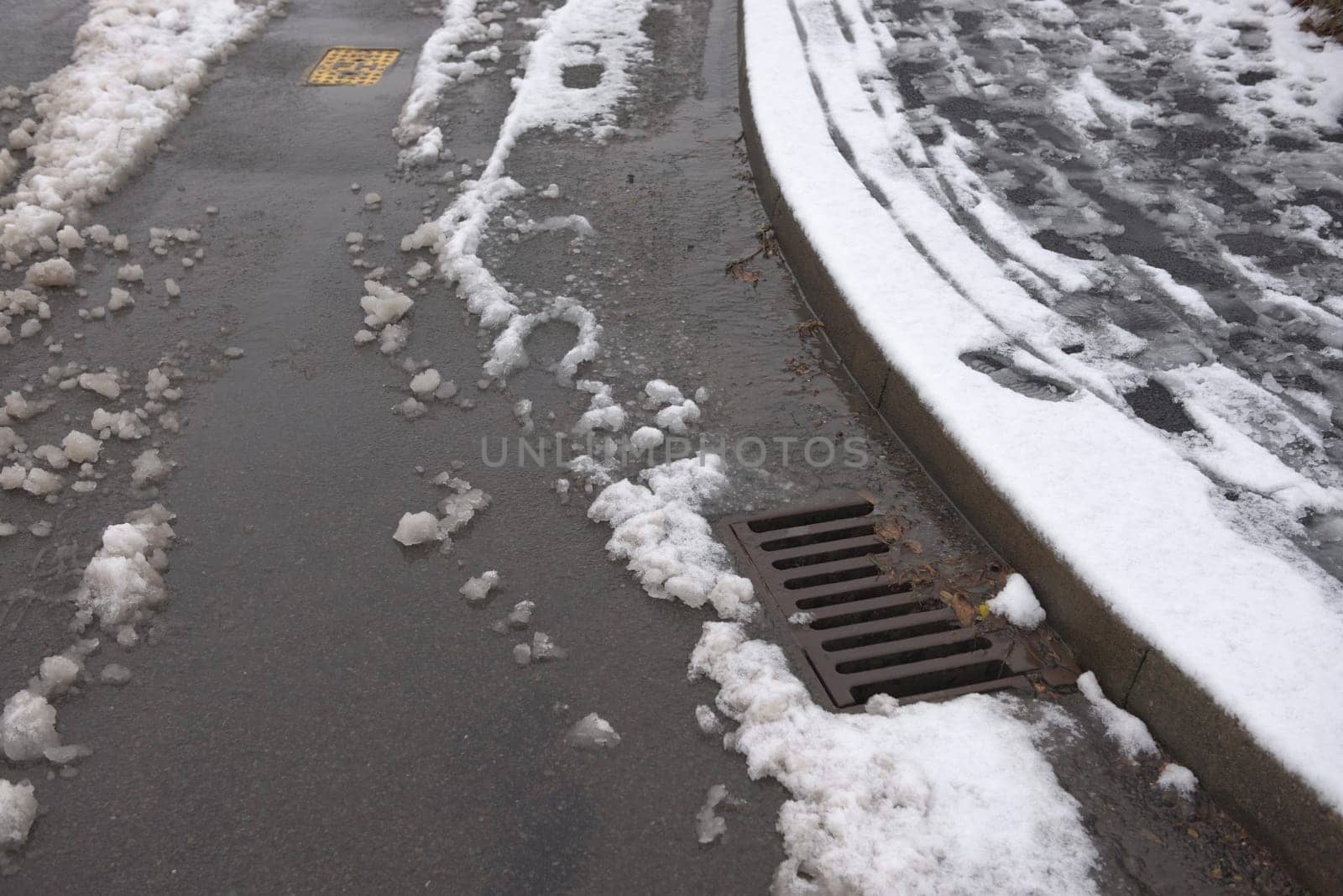 8 - Surface drain on the corner of a tarmac road, surrounded by melting snowfall. Winter environment in the UK, and water management infrastructure.