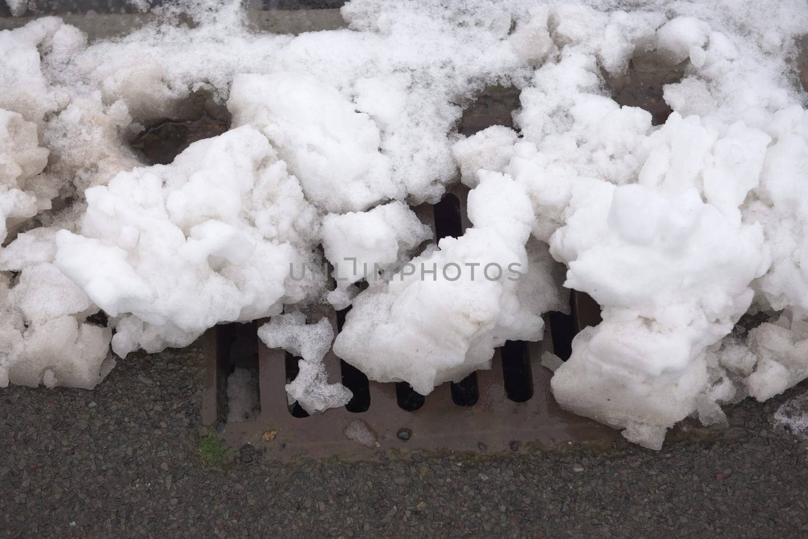 10 - Winter scene of a roadside drain covered in heavy chunks of white snow. The metal grill of the drain is nearly fully blocked by the snowfall.