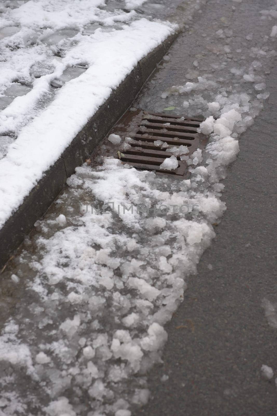 6 - Thawing snow flows into a metal curbside drain on the surface of a tarmac road. Track visible where a car has driven through the snow.